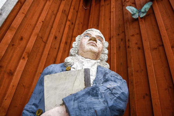 Wooden statue of botanist and founder of the binary nomenclature Carl von Linné with white hair and blue jacket in front of a wooden wall, Linnés Råshult, Älmhult, Kronobergs län, Småland, Sweden