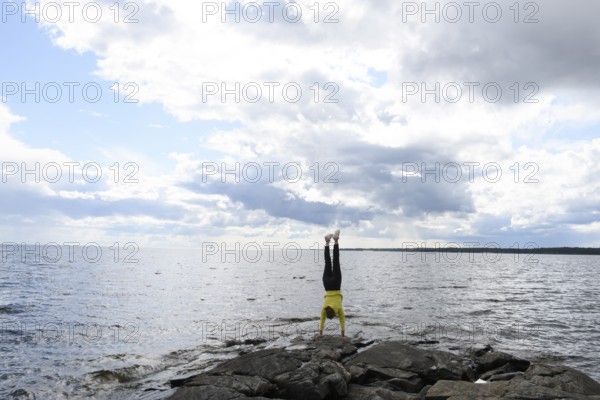 Young woman girl doing a handstand on rocks at Lake Vänern under cloudy sky, Värmlands län, Sweden