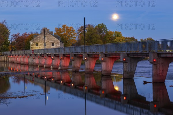 Illuminated Moulin Neuf water flow control dam and walkway over Des Mille-Iles river plus New Mill on Ile des Moulins in autumn at dusk, Old Terrebonne, Quebec, Canada