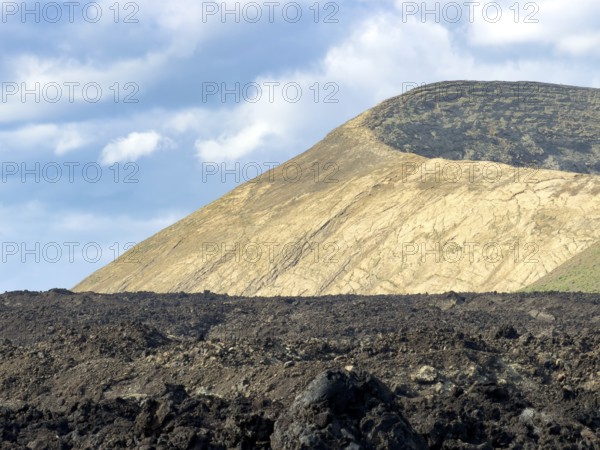 Hiking trail through lava rock to Caldera Blanca volcano against blue sky with white clouds, Mancha Blanca, Lanzarote, Canary Islands, Spain