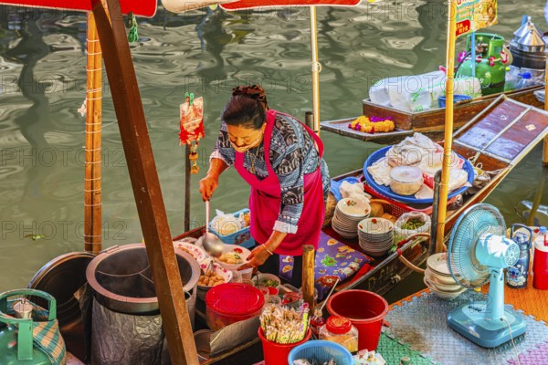 Elderly woman wearing mask offers food on her wooden boat, Damneon Saduak floating markets channel, near Bangkok, Ratchaburi district, Thailand