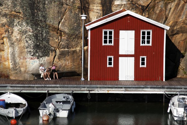 Red boathouse in front of rocks with anchored boats in calm water, Smögen, Västra Götaland, Sweden