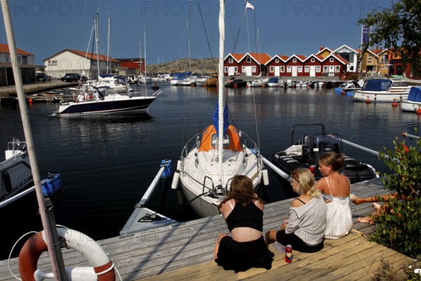 Children sit at Klädesholmen harbour looking at sailboats under sunny sky, Klädesholmen, Sweden