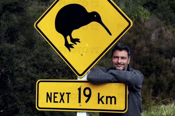 Warning sign with kiwi symbol on a road with a man in the background, Franz Josef Glacier, New Zealand