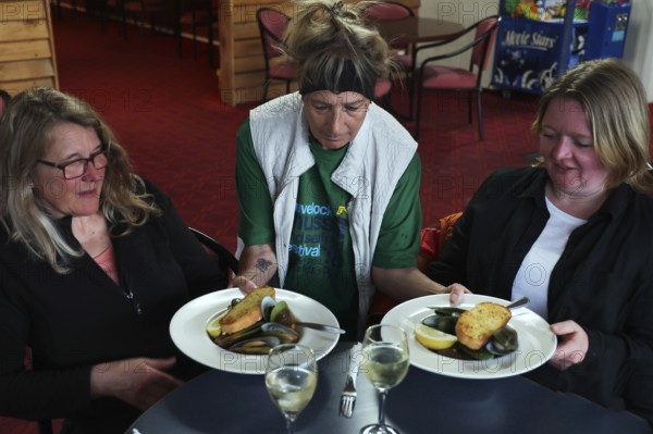 Two woman are served plates of mussels at a restaurant table by a waitress, Havelock Hotel, Havelock, New Zealand