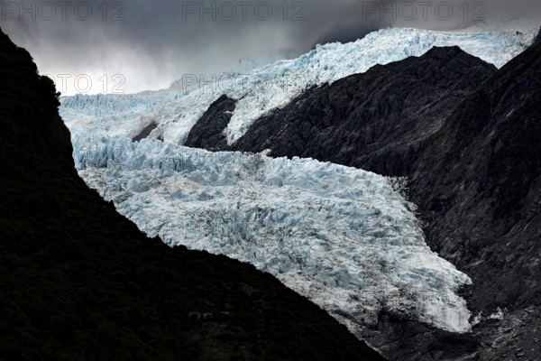 The Franz Josef Glacier contrasts strongly with dark, steep rocks under a threatening sky, Franz Josef, West Coast, New Zealand