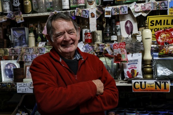 Man leans at the well-stocked bar of the Barrow Creek Hotel and smiles surrounded by souvenirs and keepsakes, Barrow Creek, Northern Territory, Australia
