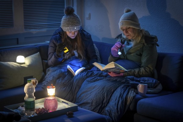 Symbolic picture blackout, extensive, prolonged power failure, in private surroundings, without electricity, heating, water, family on the sofa with warm winter clothes, sleeping bag, light from battery lights, battery lights, flashlights