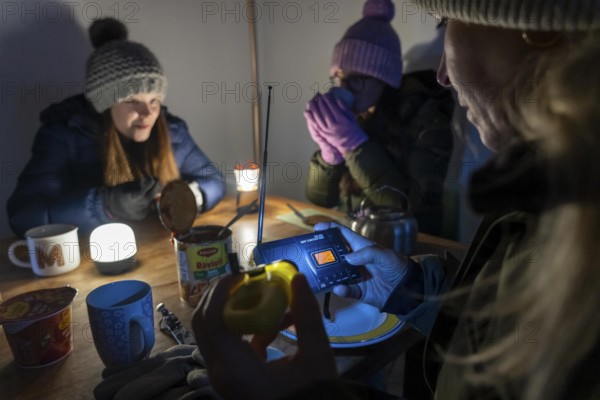 Symbolic blackout, extensive, prolonged power failure, in private surroundings, without electricity, heating, water, family at the dining table with warm winter clothes, sleeping bag, battery-operated radio, only source of information, no TV, Internet, mobile phone, light from battery lights, flashlights, food from cans, ready meals, heated on the camping stove, limited supply of food