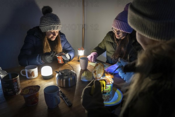 Symbolic blackout, extensive, prolonged power failure, in private surroundings, without electricity, heating, water, family at the dining table with warm winter clothes, sleeping bag, light from battery lights, flashlights, canned food, ready meals, heated on the camping stove, limited supply of food