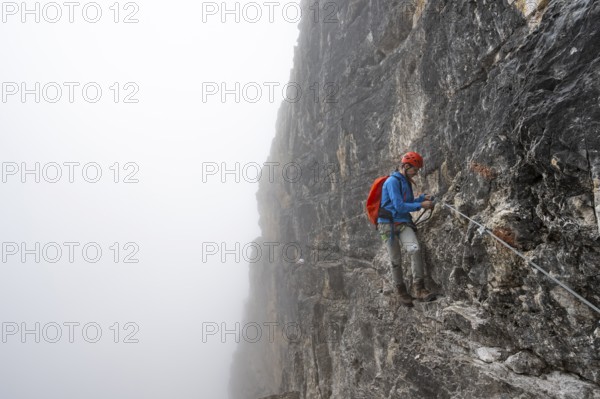 Mountaineers on a steep rock face on the Via Ferrata Oliva Detassis via ferrata in fog, steep mountains covered in clouds, Brenta Mountains, Trentino, Italy