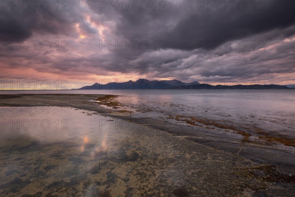 The Vestfjord in Nordland, Norway, offers a dramatic atmosphere near Bodø. Dark clouds are illuminated by the setting sun in shades of red and yellow. The rugged cliffs and mountains are reflected in the quiet fjord, which offers a breathtaking view of Lofoten
