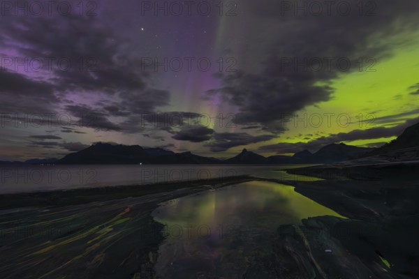 The Vestfjord in Nordland, Norway, has an enchanting atmosphere at night. Dark clouds and vivid aurora, polar lights dance over the fjord. The rugged cliffs and majestic mountains are reflected in the calm water and create a mystical backdrop