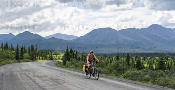 Young woman riding a bicycle on a dirt road through the tundra, mountainous landscape, Denali Park Road, Denali National Park and Preserve, Alaska, USA