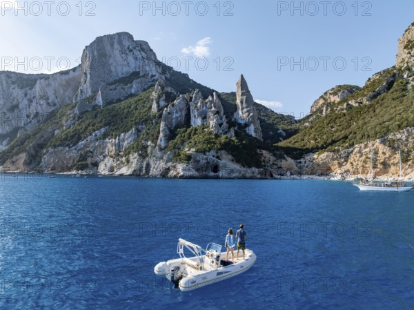 Couple on motorboat off picturesque rocky coast, cliffs with L'Aguglia pinnacle, blue sea and Cala Goloritzé beach, aerial view, Golfo di Orosei, Baunei, Sardinia, Italy