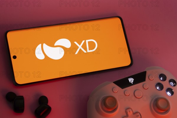 Dhaka, Bangladesh- 22 Mar 2025: Smartphone with XD logo, game controller, and earbuds on a pink background