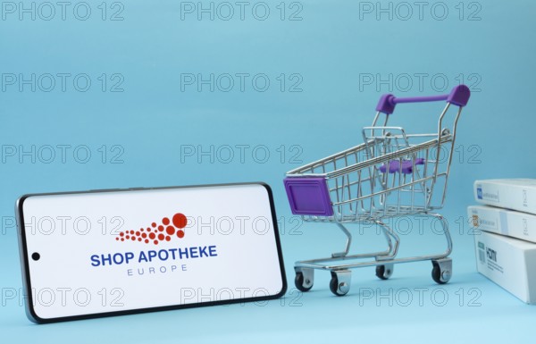 Dhaka, Bangladesh- 21 Nov 2025: Smartphone with Shop Apotheke logo next to a small shopping cart and boxes against a blue background