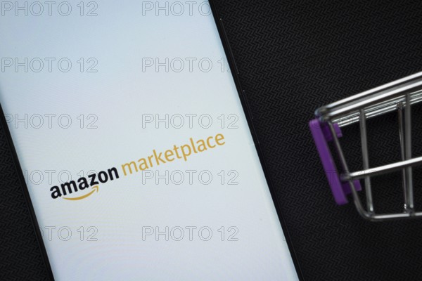 Dhaka, Bangladesh- 21 Nov 2025: A smartphone displaying the amazon marketplace logo next to a miniature shopping cart on a background