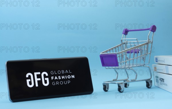 Dhaka, Bangladesh- 21 Nov 2025: Smartphone with Global Fashion Group logo next to a small shopping cart and boxes against a blue background