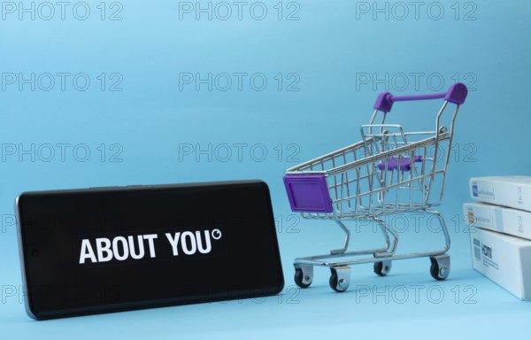 Dhaka, Bangladesh- 21 Nov 2025: Smartphone with About You logo next to a small shopping cart and boxes against a blue background