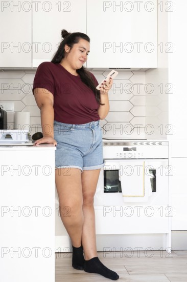 A curvy woman in a casual burgundy top and denim shorts is leaning against a kitchen counter, engaged with her smartphone in a bright, modern kitchen setting