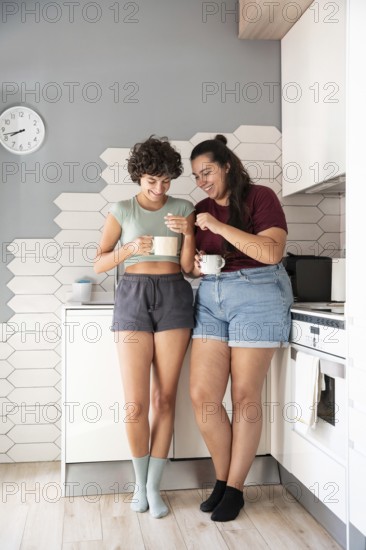 A happy lesbian couple stands in a kitchen, chatting and sharing a morning coffee. They enjoy quality time, dressed casually in shorts and t-shirts, embracing a relaxed, cozy atmosphere