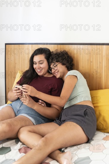 Two lesbian women sitting on a bed, enjoying a relaxed moment with a smartphone and a notebook. They are engaged and smiling, sharing a cozy and intimate atmosphere