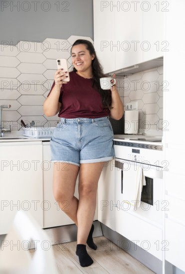 A curvy woman in casual clothes stands in a bright kitchen, holding a phone and a mug, smiling. The setting features white cabinets and a geometric tile backsplash