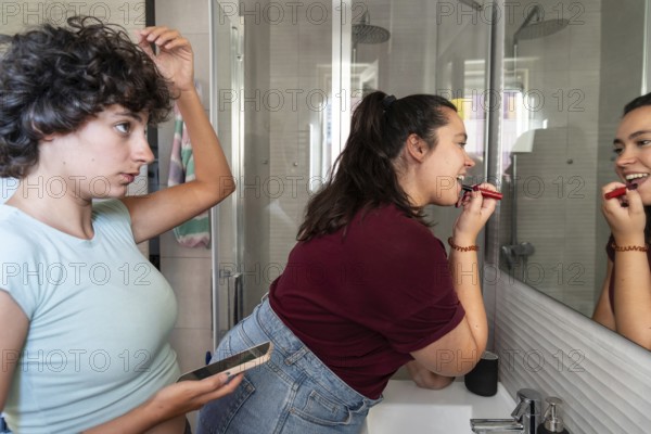 A lesbian couple shares a morning routine in the bathroom, with one applying makeup and the other styling hair, creating a candid and intimate moment