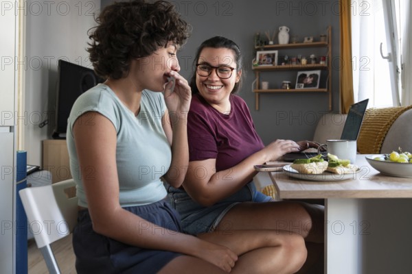 A lesbian couple shares a cozy breakfast in their modern kitchen, with one partner working on a laptop. The warm sunlight and smiles create a relaxed, cheerful atmosphere