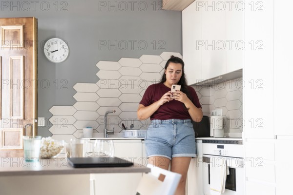 A curvy woman in a casual outfit stands in a modern kitchen, intently using her smartphone. The stylish kitchen features hexagonal tiles and a clock