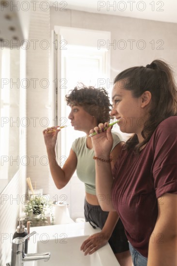 A lesbian couple shares a morning routine in the bathroom, with one applying makeup and the other styling hair, creating a candid and intimate moment