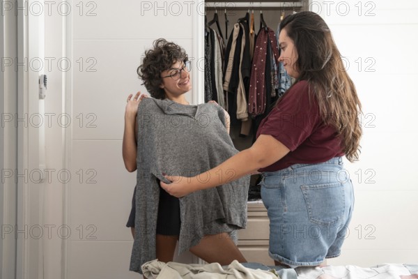 A joyful lesbian couple shares a moment while picking out clothes. One holds up a gray sweater, seeking the other's opinion, highlighting domestic harmony and love
