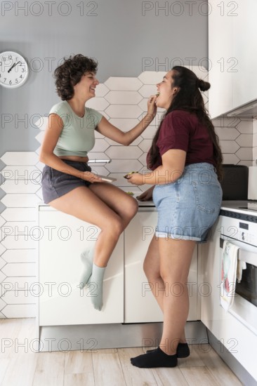 A happy lesbian couple shares a playful moment in a modern kitchen. One partner sits on the counter, feeding the other with a smile, showcasing a warm, intimate connection