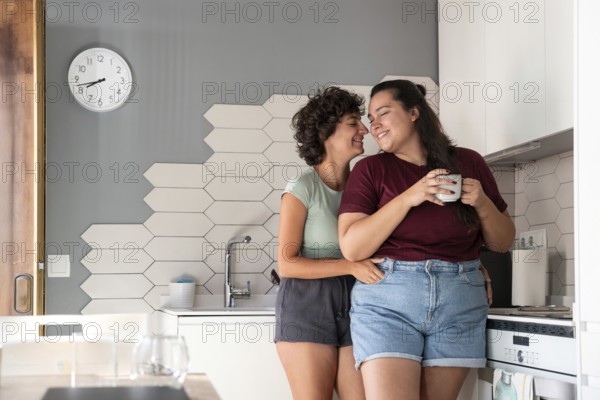 A happy lesbian couple shares a playful moment in a modern kitchen. One partner sits on the counter, feeding the other with a smile, showcasing a warm, intimate connection