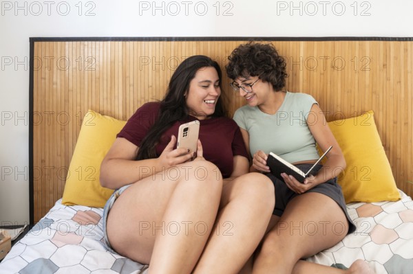 Two lesbian women sitting on a bed, enjoying a relaxed moment with a smartphone and a notebook. They are engaged and smiling, sharing a cozy and intimate atmosphere