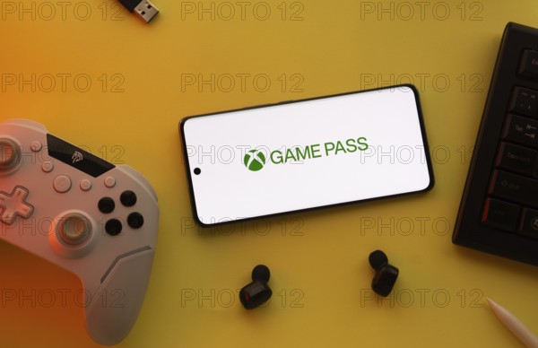 Dhaka, Bangladesh- 15 July 2025: Smartphone with Xbox game pass logo, game controller, and earbuds on a yellow background