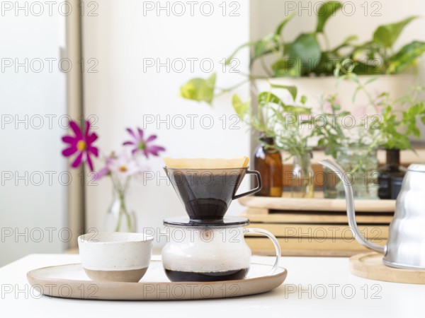 Brewing pour over coffee with a modern kettle, paper filter, and glass carafe, with plants and flowers adding a touch of nature