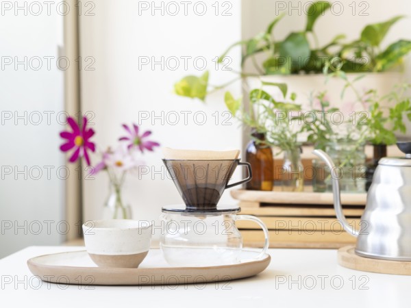 Modern pour over coffee brewing setup sits on a white table, with houseplants and flowers adding a touch of nature to the scene