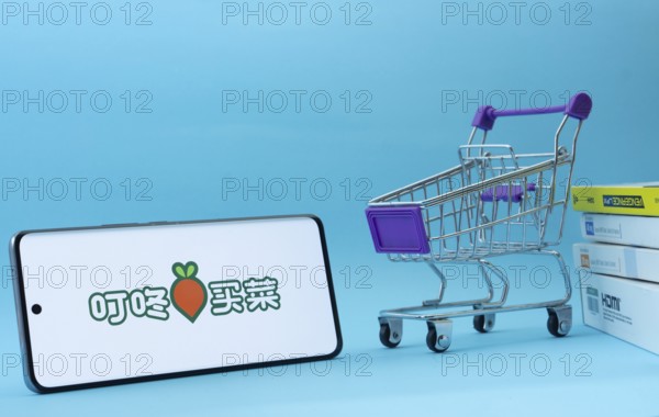 Dhaka, Bangladesh- 03 July 2025: Smartphone with Dingdong Maicai logo next to a small shopping cart and boxes against a blue background