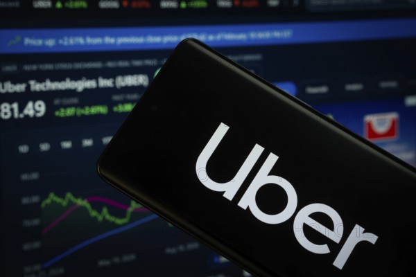 Dhaka, Bangladesh- 15 July 2025: Uber logo is displayed on a smartphone screen, with a stock graph webpage visible on a computer screen in the background