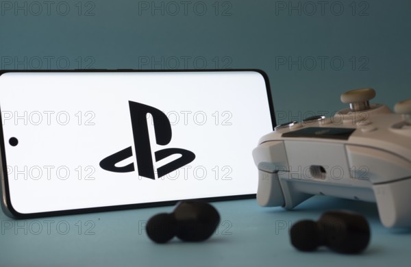 Dhaka, Bangladesh- 26 July 2025: A smartphone with a PlayStation icon next to a controller and earbuds on a blue surface