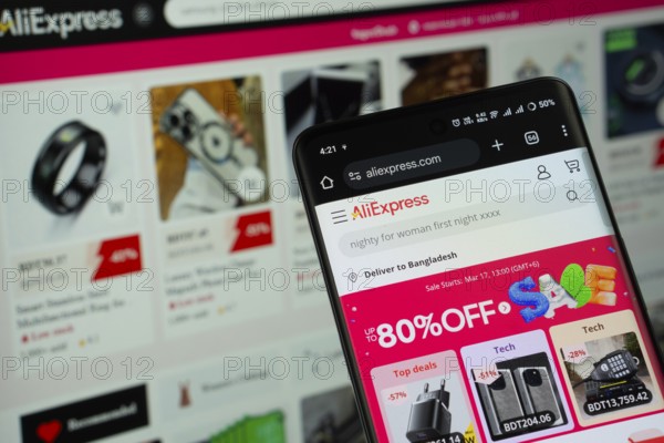 Dhaka, Bangladesh- 05 March 2025: AliExpress logo is displayed on a smartphone screen, with the website is visible on a computer screen in the background