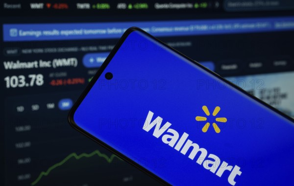 Dhaka, Bangladesh- 15 July 2025: Walmart logo is displayed on a smartphone screen, with a stock graph webpage visible on a computer screen in the background