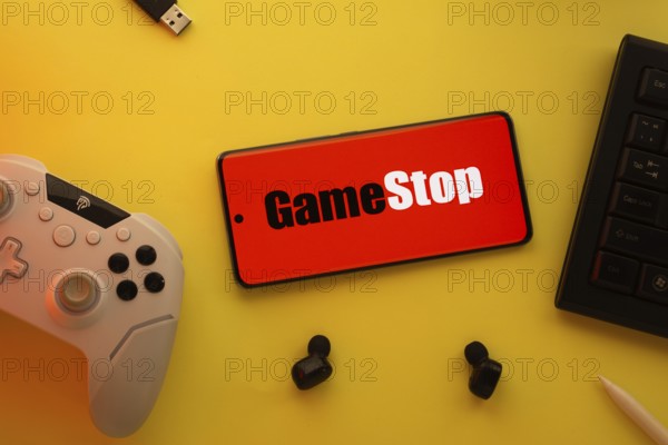 Dhaka, Bangladesh- 15 July 2025: Smartphone with GameStop logo, game controller, and earbuds on a yellow background