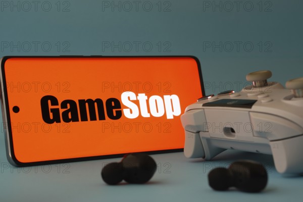 Dhaka, Bangladesh- 15 July 2025: A smartphone with a GameStop icon next to a controller and earbuds on a blue surface