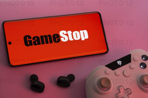 Dhaka, Bangladesh- 15 July 2025: Smartphone with GameStop logo, game controller, and earbuds on a pink background