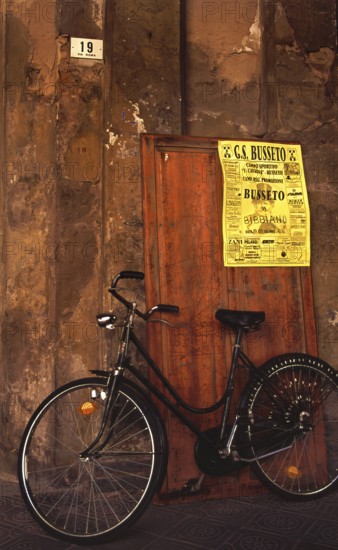 A bike stands in front of an old house wall with a soccer poster of GS Busseto, in honor of Giuseppe Verdi, Via Roma 19, Busseto, Province of Parma, Emilia-Romagna, Italy