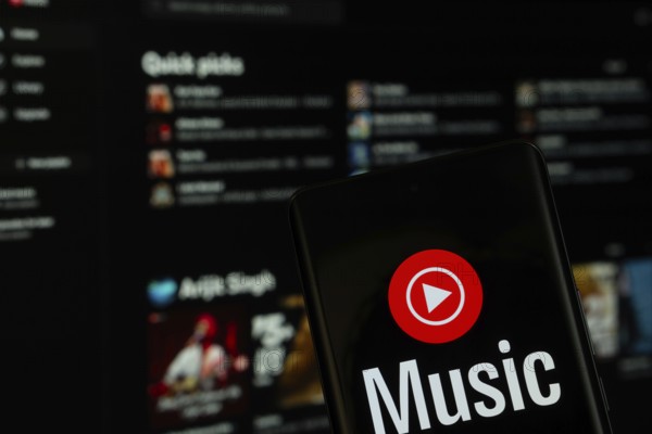 Dhaka, Bangladesh- 04 July 2025: Youtube Music logo is displayed on a smartphone screen, with the website visible on a computer screen in the background