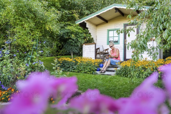 A woman sits relaxed on the small veranda of a garden shed and reads. Blooming summer flowers in the garden. Germany
